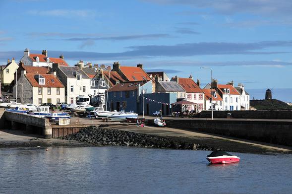 St Monans. The image shows a view over a harbour to a slipway with, beyond it, a collection of red tile roofed houses. There are numerous small boats parked at the top of the slipway. A windmill can be seen in the distance on the right. The harbour is in sunlight.