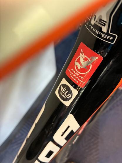 Close-up of a bicycle frame featuring stickers, including a red and white AUDAX UK sticker for long-distance cycling and a round Vélo Culture logo. The frame is primarily black with orange accents.