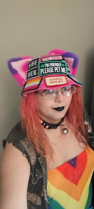 selfie of vanta rainbow black in her iconic hot pink bucket hat with cat ears and patches sewn on and lesbian collar and black lipstick and purple eyeliner and a rainbow top. she is smiling