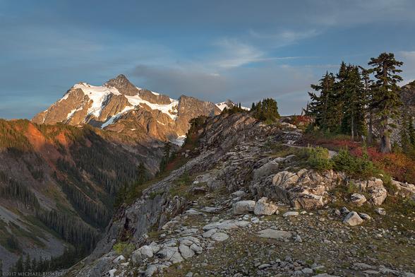 A photo from the edge of a ridge. On the left the rocky ground drops off dramatically, on the right the rocky ridge has some small foliage from blueberry bushes and a few conifer trees.  Evening light shines on a mountain peak in the left rear of the photograph.  Most of the rest of the photo is in shade.