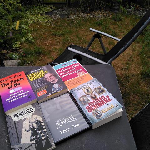 Ein Gartentisch im Garten zeigt fünf Bücher und eine DVD, die thematisch um Wissenschaft, Skepsis und kritisches Denken kreisen. Zu sehen sind unter anderem „The Power of No“ von Dana Buchzik, zwei Werke von Jens Foell, „The Hoax-Files“, „Hoaxilla – Year One“ und der Krimi „Kohlrabenschwarz“. Daneben steht ein schwarzer Gartenstuhl, im Hintergrund ist eine grüne, leicht verwilderte Wiese mit Sträuchern zu erkennen.