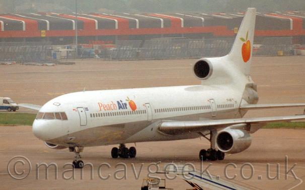 Side view of a 3 engined jet airliner taxiing from right to left.
The plane is mostly white, with a silver belly.
There are orange and blue "Peach Air" titles on the upper forward fuselage, next to an image of an orange coloured fruit, this logo also appearing in a larger form on the tail.
Grey concrete apron fills most of the frame, with red, white, and brown cargo sheds stretching across the image in the background.