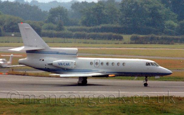 Side view of a 3 engined BizJet moving from left to right.
The plane is mostly white, with 3 thin blue stripes running along the lower body, and another 3 stripes running along the bottom of the centre engine air intake and tail.
The blue registration "VR-CAE" is on the side of the engine pods mounted on the sides of the rear fuselage.
Green grass fills the foreground, with more grass and taxiways in the background leading up to trees in the distance.