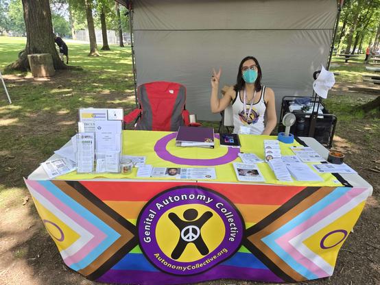 A table covered in genital autonomy literature with an intersex flag as a table top, and the Genital Autonomy Collective banner in front.  Alliana of GAC sitting on the right side next to a red chair under a canopy making a peace sign.