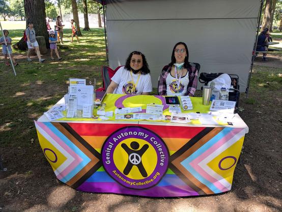 A table covered in genital autonomy literature with an intersex flag as a table top, and the Genital Autonomy Collective banner in front.  Alliana of GAC sitting on the right side next to Perry of GAC.