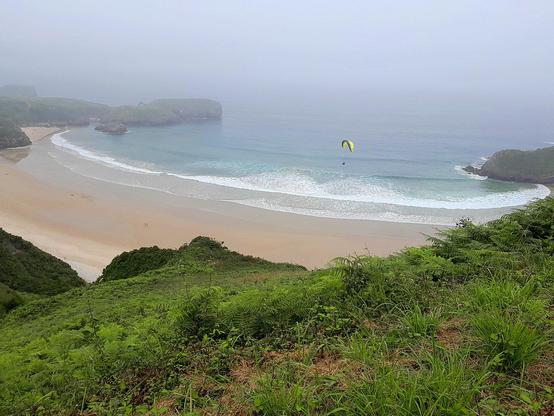 Playa en forma de concha vista desde el verde anfiteatro que la cierra, de clara arena tostada y azules aguas turquesa, las ondas de las olas forman cenefas blancas diluyendo el límite de tierra y mar. El horizonte difuminado cualquier atisbo de frontera entre cielo y mar. Se trata de una conocida playa llanisca, pionera del nudismo ideológico, que el otro siempre se practicó.