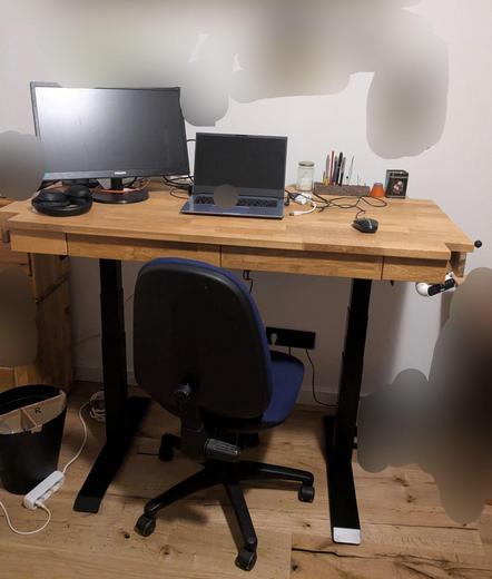 Holztischplatte mit Schubladen, auf dem Gestell eines Stehschreibtisches in Stehhöhe. Auf dem Tisch ein Laptop, Monitor, Kopfhörer, Stiftehalter, Maus.