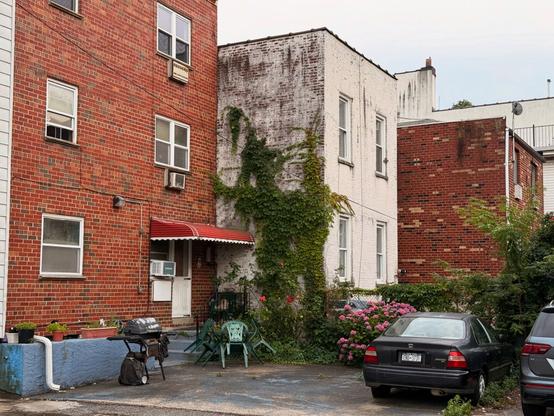 A grey sky with a brick building with a red awning with a white stripe. Green vines grow next door on a white painting building. A black bbq grill sits on the back driveway in the foreground. A black car sits off to the side.