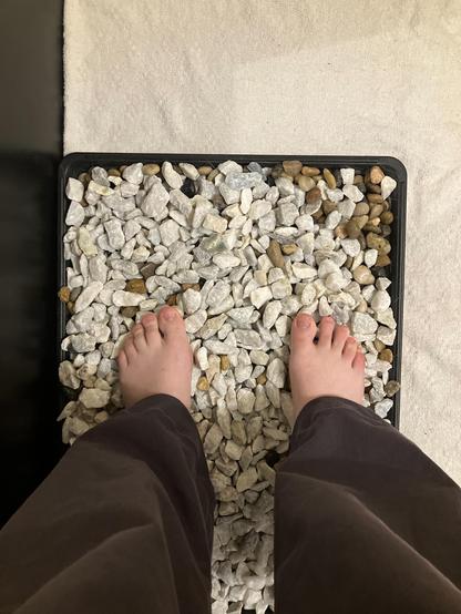 Feet in a rock garden from the perspective of the owner looking down over legs clad in a pair of baggy pants. White feet standing in a tray of stones, The tray is on top of a white towel.