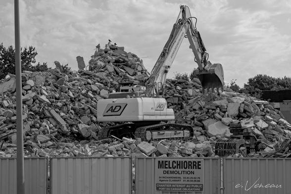 A mechanical shovel stands on a pile of rubble, the last vestige of the tower block. In front of it is a works barrier bearing the name of the demolition company.