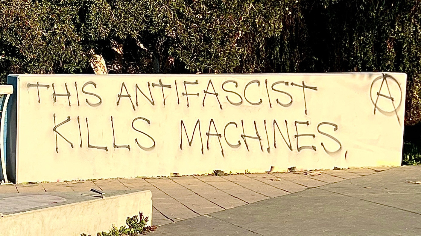 The image shows a concrete wall with graffiti written on it. The graffiti reads "THIS ANTIFASCIST KILLS MACHINES," with the word "ANTIFASCIST" and the phrase "KILLS MACHINES" in capital letters. To the right of the text, there is a symbol resembling an anarchy symbol, which is an "A" inside a circle. The wall is light-colored, and the background includes some greenery, suggesting an outdoor setting. The ground in front of the wall is paved with square tiles. The text is written in a rough, hand-drawn style, indicating it was likely spray-painted or written with a marker.