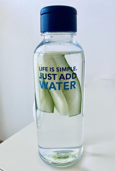 Eine klare Wasserflasche, gefüllt mit Wasser und Gurkenstreifen, mit dem Text "LIFE IS SIMPLE. JUST ADD WATER." auf der Vorderseite. Die Flasche hat einen blauen Deckel und steht vor einem hellen Hintergrund.