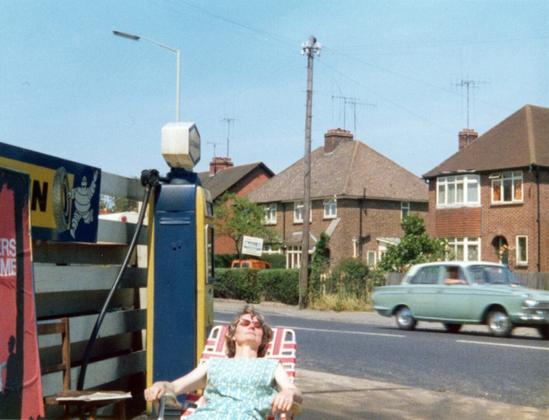 With red brick houses and Basingstoke Road (complete with passing car) behind her, Mary is sunbathing in a red and white lounging chair next to a blue and yellow petrol pump. Next to that a sign for Michelin tyres is partially visible.