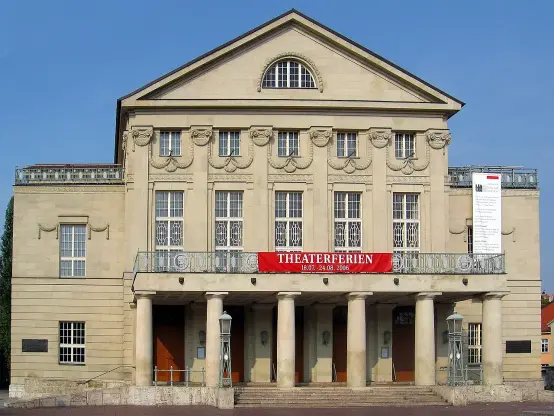 The image depicts a building that appears to be a theater, characterized by its classical architectural style. The facade features a prominent entrance with columns, decorative elements, and a balcony. A banner mentioning "THEATERFERIEN" is displayed prominently, indicating some kind of event or festival. The sky is clear, suggesting a bright day.

Image Credits: Wikimedia / Andreas Trepte / CC BY-SA 2.5