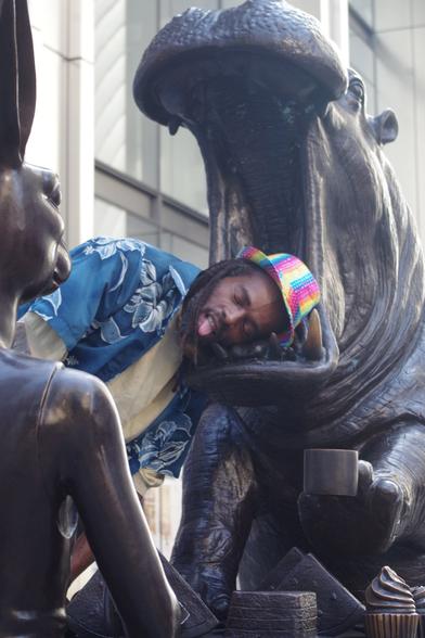 A man putting his head sideways against the mouth of a sculpture of a creature with a hippo's head and a human body, with one hand holding coffee and cupcakes in front of the torso. In the foreground is a creature with a dog's head and a human body, as part of the artwork *The White Rhino Was Thinking Ahead with Dogman's Encouragement*.