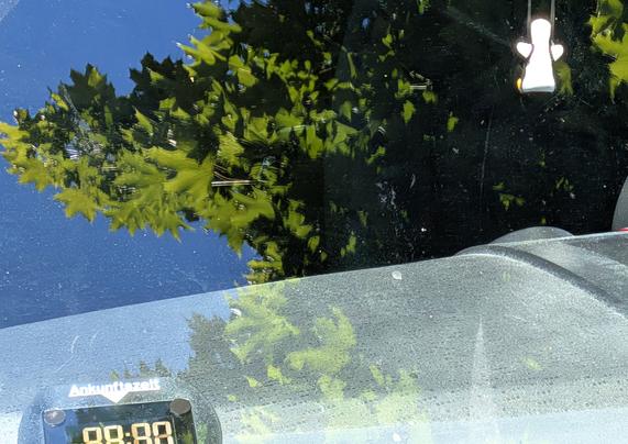 Dieses Hochformat-Foto, von außerhalb des Autos auf die Windschutzscheibe gerichtet, zeigt ein faszinierendes Spiel von Reflexion und Durchblick.

Oben spiegelt sich ein Baum mit leuchtend grünen Blättern und Ästen im tiefblauen Himmel der Scheibe. Diese spiegelnde Oberfläche fängt die Natur in idealisierter Klarheit ein, fast wie ein Gemälde.

Darunter, durch die leicht verschmutzte Scheibe, erscheint die gedämpfte Realität. Die Textur des Glases mit Staub und Schlieren dient als Filter, der die Umgebung in weichere, dunklere Töne taucht. Die Baumumrisse verschwimmen diffus, was die Komplexität der Wahrnehmung betont.

Unten links ist ein digitales Display im Auto sichtbar, das "Ankunftszeit" und "88:88" anzeigt. Dieses Detail verankert das Bild in der Moderne und kontrastiert mit der organischen Reflexion.

Ein Blickfang ist eine kleine, helle, stilisierte Figur, die im Fahrzeuginneren hängt. Sie fängt das Licht ein und wirkt fast ätherisch, was dem Bild eine persönliche, schützende Dimension verleiht.

Die Komposition spielt meisterhaft mit Schein und Realität. Die Windschutzscheibe verbindet und trennt zwei Welten: die spiegelnde Oberfläche und den Blick ins Innere. Die lebendige Reflexion gegenüber der gedämpften Transparenz erzeugt visuelle Spannung. Die hängende Figur ist ein stiller Zeuge dieses Dialogs, der zum Nachdenken über Wahrnehmung und Schönheit einlädt.