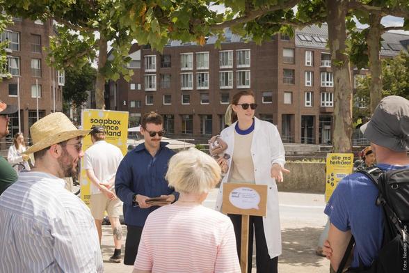 Soapbox Science Vortrag mit einer Person in einem Laborkittel, die vor einem Publikum spricht. Ein Banner mit der Aufschrift 'soapbox science rheinland' ist zu sehen.