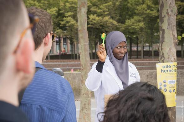 Eine Person im Laborkittel präsentiert etwas beim Soapbox Science Event, während das Publikum zuschaut. Ein Plakat ist im Hintergrund sichtbar.