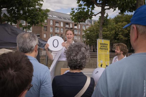 Vortrag bei Soapbox Science Event, eine Person zeigt ein rundes Zeichen mit der Zahl '0'. Mehrere Zuschauer sind im Vordergrund.