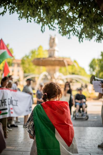 Une femme de dos avec un drapeaux palestinien sur les épaules. Devant elle, la manif et la Rotonde d'Aix