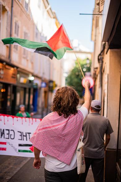 Un manifestant avec un kéfié rouge, qui brandie un drapeau palestinien, juste derrière la banderole de tête