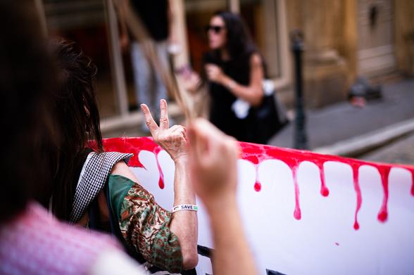 Une main tenant la banderole de tête en faisant un V avec les doigts. La banderole est notamment blanche avec des trace de peinture rouge laissant penser à du sang qui à coulé.