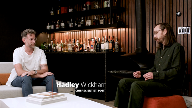 Hadley Wickham in a white t-shirt and jeans sits on a light-colored couch across from Michael Chow, wearing a dark green shirt and pants, who is seated on an orange pouf. Between them is a white coffee table with books. In the background, there's a dark bar with shelves displaying numerous bottles, a wood-paneled wall, and a black, modern fireplace. Text overlay reads "Hadley Wickham CHIEF SCIENTIST, POSIT".