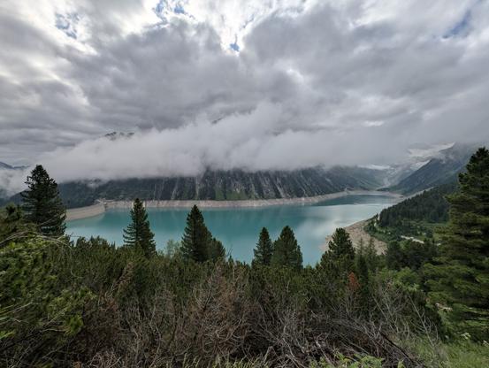 The image showcases the stunning Schlegeisspeicher at the end of the Zillertal in Austria. The lake exhibits a vibrant turquoise hue, beautifully framed by towering mountains, which are enveloped in clouds. To the left, a dam is visible. In the foreground, green vegetation contrasts with dry branches. The sky is overcast, with heavy clouds casting a moody atmosphere, while a distant glacier can be seen on the far side of the lake.