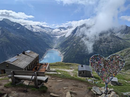 The image presents a breathtaking mountain landscape taken right above the Olperer hut, which is visible on the left. In the foreground, a wooden bench provides a resting spot, with trekking poles leaning against its backrest, suggesting recent activity. To the right, a heart-shaped structure adorned with love locks adds a romantic touch. The background showcases majestic snow-capped mountains under a partly cloudy sky and the turquoise Schlegeisspeicher in the valley below completes this idyllic alpine vista.