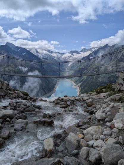 Stunning view taken near the Olperer hut, featuring a suspension bridge that is popular on Instagram. From a rocky vantage point, a stream flows through the foreground, leading the eye towards the bridge. In the middle ground, the Schlegeisspeicher is nestled between mountains, which display patches of snow and greenery. The sky above is a vibrant blue, dotted with scattered white clouds.