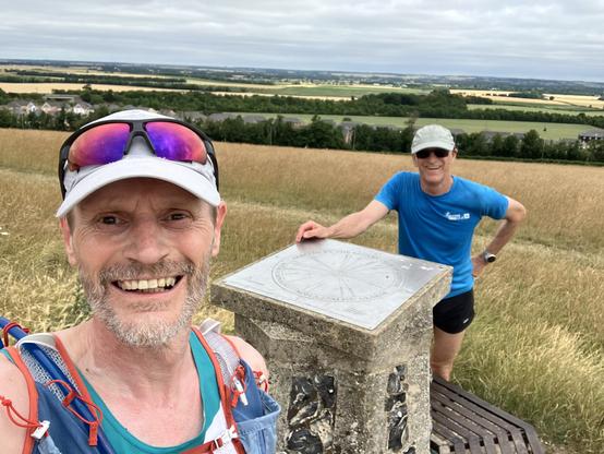 out on the hills at the weekend with New from C&C running club. two runners, nearest one smiling a toothy grin wearing a white hat and sunglasses, in a running vest, at a trig point on Royston Heath. New in th background also wearing a hat and shades, and a blue t shirt. background is the grasss slopes of the heath giving way to farmland and trees in the distance under an overcast sky