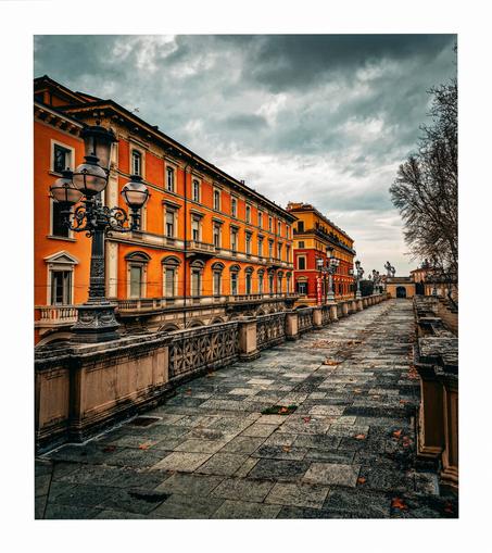 This photo is of a long, paved walkway, a terrace, lined with ornate stone railings.  The walkway has a row of tall, vibrant orange buildings with classical architecture on its left, under a dramatic, overcast sky.  Scattered leaves are visible on the path, and bare trees are present in the background. (It really early spring)  The overall mood is quite dark and moody.