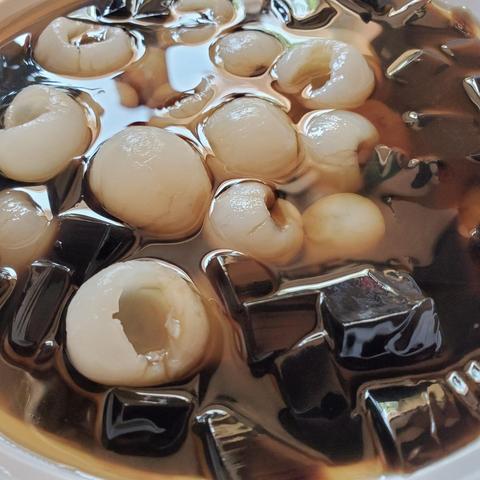 High-angle, close-up shot. Dominant elements are small, light beige spheres (lychees) and dark-brown cubes (grass jelly) suspended in a light-brown liquid. The shot is tightly framed, minimizing background. Lighting creates highlights on the gelatin and liquid surfaces, enhancing texture. The composition is dense and textural, emphasizing the ingredients' contrast.