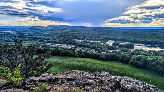 A soft blue sky with a mass of clouds passing a few miles to our west, dropping some visible rain showers on the distant mountains. We are following the western edge of an open stretch of ridge crest covered with grass, scrubby plants and brownish gray rock formations. Several hundred feet below is a broad green valley with lots of forest and scattered clusters of houses and other buildings. A large rolling meadow is below us, with a built-up strip of larger buildings just beyond. The corner of a large lake is on the right not far away. On the far side of the valley, several miles away, a long traprock ridge runs along the horizon, culminating in a pair of south-facing cliffs. A portion of that ridge is catching the rain showers.