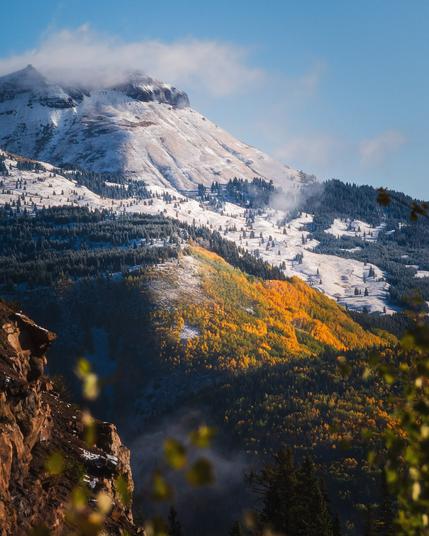 A scenic mountain landscape featuring a snow-capped peak in the background, with lower slopes covered in a mix of evergreen trees and vibrant autumn foliage in shades of yellow and orange. The sky is clear and blue, and there are clouds clinging to the snow-capped peak.
