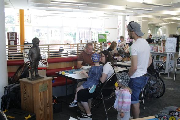 Part of Barry library with tables and chairs laid out and people sat at them fixing things. In the foreground at the nearest table stands a family. An adult sat at a table with a small boy on their lap while another adult stands behind with a small girl standing beside them eating a cupcake.