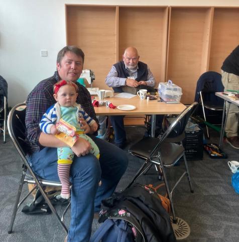 A person with light skin tone and short brown hair sitting at table with a very young child wearing a red bow in their hair on their knee. Behind them Peter, a bald man with light skin tone with a tan sits at a table sewing something up. On the table are also mugs and a plate and other sewing equipment.