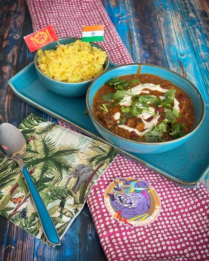 Das Bild zeigt eine appetitliche Mahlzeit auf einem blauen Holztisch. Im Vordergrund befindet sich ein blauer Teller mit einer Schüssel voller Curry, das mit frischem Koriander und einer weißen Sauce garniert ist. Neben dem Curry ist eine Schüssel mit gelbem Reis, der mit einem kleinen indischen Flaggenstäbchen und einem veganen Symbol garniert ist. Ein silberner Löffel liegt auf einem bunten Tuch mit tropischen Motiven, das eine Elefantendarstellung zeigt. Ein rosa Tuch mit weißen Punkten liegt unter dem Teller. Ein kleiner Aufkleber mit einem Vogel und dem Text "BECOME UNGOVERNABLE" ist ebenfalls sichtbar. Die Farben und die Präsentation der Mahlzeit vermitteln ein fröhliches und einladendes Ambiente.