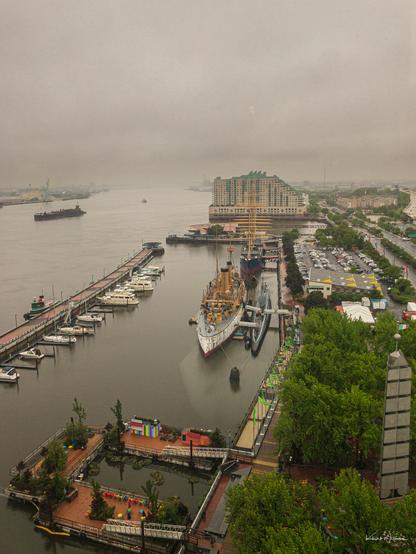 A foggy riverside scene showing a historic warship and submarine docked at a marina in front of a large hotel complex. The water is calm under overcast skies, with scattered boats and barges visible in the distance. A tree-lined promenade and a small public park with colourful umbrellas and a pond sit in the foreground. The atmosphere is quiet and muted by the low grey clouds.