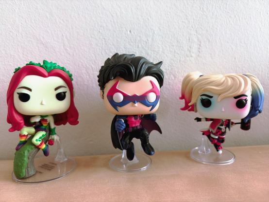 Three Funko Pop figures displayed on a light wooden surface against a textured white wall. From left to right: Poison Ivy with vibrant red hair and green leaf-adorned Progress Pride Flag outfit holding a plant stem, Robin in a Bisexual Pride Flag themed outfit, and Harley Quinn with blonde hair styled in pigtails wearing a Bisexual Pride Flag themed outfit. All figures are positioned on clear plastic stands, creating a dynamic superhero trio showcase with Batman universe characters.​​​​​​​​​​​​​​​​