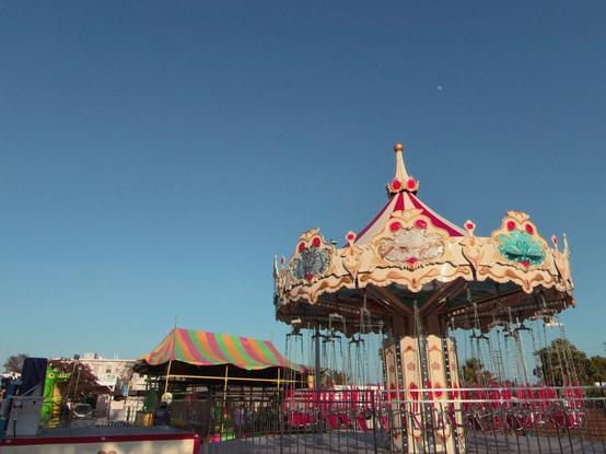A vintage carousel with ornate cream-colored canopy featuring decorative red and teal painted details sits prominently in the foreground against a clear blue sky. The merry-go-round’s elaborate top is adorned with classical carnival motifs and a golden finial. Pink safety barriers surround the ride’s base. To the left, a colorful striped circus tent in rainbow hues adds vibrant contrast to the scene.