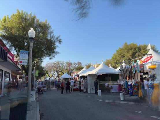 A bustling outdoor street festival with white canvas vendor tents lining both sides of a closed street. People browse merchandise displays hanging from tent frames while others walk leisurely through the scene. Mature trees with full green foliage provide natural shade, contrasting against a clear blue sky. On the left, storefronts including a gelato shop are visible, with decorative street lamps adding urban charm. The atmosphere appears relaxed and sunny, typical of a weekend community market or arts festival. Colorful banners and merchandise create vibrant pops of color throughout the scene.​​​​​​​​​​​​​​​​