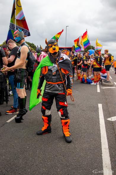 A person (it/its) amongst a crowd in a pride parade on an overcast day. It's wearing an orange pup hood, full orange Motocross gear, a paracord collar/leash, and an Aromantic flag hung from its neck. Amongst the crowd is a rugby team and other pups in various kink gear. Various flags are being flown, including one with the @nuzzles.uk breasted across a LGBTQIA+ flag.