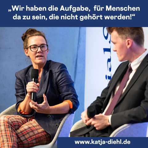 Katja Diehl spricht in einem Podiumsgespräch engagiert in ein Mikrofon, neben ihr sitzt ein junger Mann im Anzug. Sie trägt einen dunkelblauen Blazer mit hochgekrempelten Ärmeln und karierte Hosen. Oben im Bild steht das Zitat: „Wir haben die Aufgabe, für Menschen da zu sein, die nicht gehört werden!“ Unten ist die Website „www.katja-diehl.de“ eingeblendet. Die Szene spielt sich auf einer Bühne im Rahmen einer Konferenz ab.