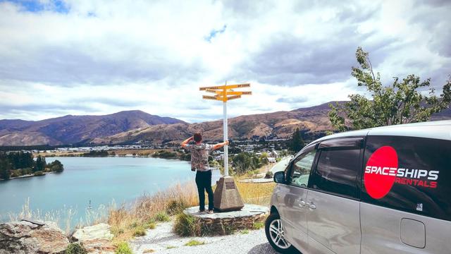 Space Traveller standing on a hill next to a sign post, enjoying the view. Lake & mountains in front of them. Spaceships campervan parked on the right. Beautiful day with white fluffy clouds.
