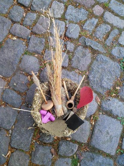 Auf grauen Pflastersteinen steht eine Grasschüssel mit Graspapier, ein glatt abgeschnittenes Grasbüschel, eine kleine Flöte, eine Tabakspfeife, eine Miura-Faltung, eine Spindel mit aufgewickelten Garn, ein Springkraut-Modell, ein Ahornsamenmodell ind Brennnesselfasern.