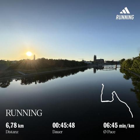 Am Horizont eine Kirche (Magdeburger Dom), im Vordergrund, ein nahezu spiegelglatter Fluss (die Elbe) links in Richtung der Stadt, die untergehende Sonne die aufgehende Sonne, der Fluss ist gesäumt von Bäumen, Büschen und Wiesen, die Stadtsilhouette von Magdeburg über die Elbe Richtung Dom geblickt am Abendhimmel