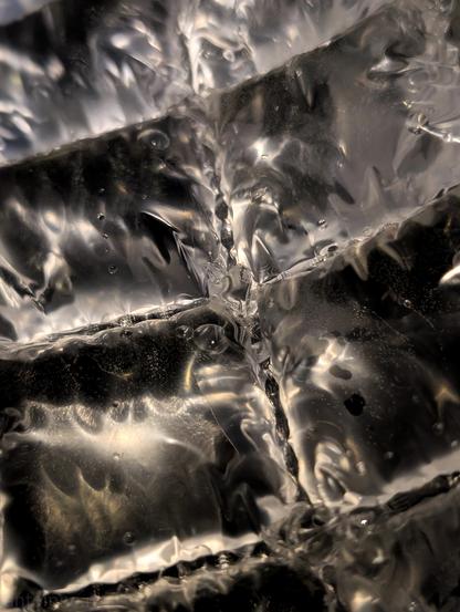 This is a close-up, abstract photograph that captures a dynamic and somewhat chaotic scene of water interacting with a textured and rippled surface, made of plastic.

The image is dominated by various shades of silver, grey, and black, with glints of light creating highlights that suggest reflectivity. There's a strong sense of movement, with swirling patterns and what look like bubbles or air pockets suspended within the liquid. Some areas are sharply defined, showing distinct lines and creases, while others are blurred, contributing to the sense of fluidity.