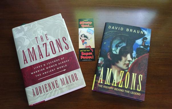 Two books on a wooden table. One book cover has a grayed out image of women in battle and dark red type (The Amazons: Lives & Legends of Women Across The Ancient World by Adrienne Mayor). The second book has an image of a helmeted woman painted in color (Amazons: The History Behind The Legend by David Braund). In between the books is a Wonder Woman book mark that reads: "Lasso a Library Card and Find Your Super Power!"