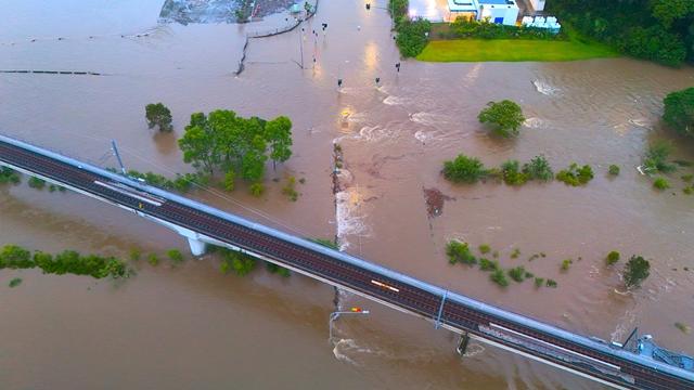 An aeriel photo showing Kedron Brook bursting its banks, enveloping a road, and the nearby site where Toombul Shopping Center used to stand. Traffic lights are standing visible within the flood waters, still functioning